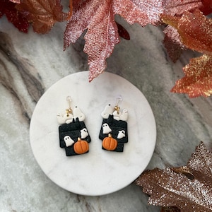 May include: A pair of drop earrings featuring a black rectangular doghouse shape with a white dog figure on top, small white ghost figures, and an orange pumpkin at the base. The earrings are attached to gold-toned metal hooks for pierced ears.