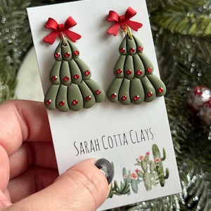 May include: A pair of olive green Christmas tree earrings featuring red rhinestone accents and a small red bow at the top. The earrings are attached to a white display card with the text Sarah Cotta Clays and a small illustration of cacti.
