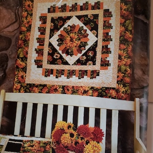 May include: A quilt block featuring a sunflower design in shades of orange, yellow, and brown. The quilt block is surrounded by a border of black and white squares. The quilt block is hanging on a white wooden bench with a basket of sunflowers in front of it.
