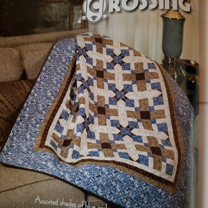 May include: A quilt with a patchwork design featuring blue and brown squares and stars. The quilt is folded on a brown couch with a floral pattern. The text "Canterbury Crossing" is visible at the top of the image.