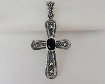 Puede incluir: Un colgante de cruz de plata con una piedra de ónix negro en el centro. La cruz está decorada con piedras de marcasita.