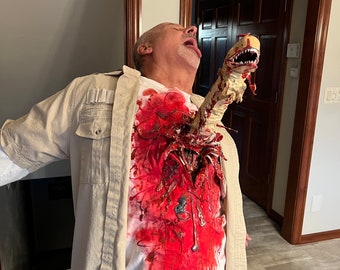 May include: A man in a white shirt and beige jacket with a fake alien creature bursting out of his chest. The creature has a long, thin body with a large head and sharp teeth. The man is looking up in pain, and there is blood splattered on his shirt.