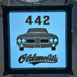 May include: A backlit sign featuring a classic car design. The sign displays the number "442" above a car silhouette and the word "Oldsmobile" in a stylized font. The frame is black, and the background is a light blue.
