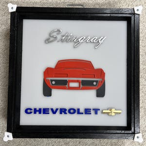 Może przedstawiać: Oprawiona grafika przedstawiająca czerwoną Chevroleta Corvette Stingray. Samochód jest przedstawiony od tyłu, z czarnymi oponami i srebrnym emblematem "Stingray" powyżej. Słowo "CHEVROLET" jest w kolorze niebieskim, z złotym logo muszki poniżej.