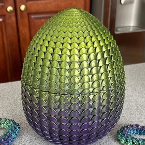 May include: A decorative egg-shaped object with a textured, scale-like pattern. The color gradient transitions from green at the top to purple at the bottom. The egg is sitting on a gray surface.