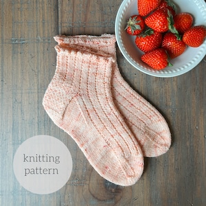 May include: A pair of pink and white knitted socks with a ribbed pattern. The socks are on a wooden surface with a bowl of strawberries in the background. The text "knitting pattern" is written on a white circle.