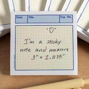 May include: A collection of sticky notes with a grid design and blue borders. The central note says "I'm a sticky note and measure 7.62cm x 7.30cm". A yellow pen and a grey marker are in the foreground. The notes are on a wooden surface.