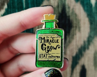 Puede incluir: Un pin de esmalte verde con forma de botella de poción. El pin presenta las palabras "Miracle Grow" y "STAT for cultures" en un estilo vintage. La botella tiene una tapa dorada y un borde plateado.