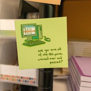 May include: A lime green sticky note with a cartoon of a computer monitor, keyboard, and mouse. The text on the note reads, "are you sure all of the QC points crossed over and passed?" The note is attached to a black metal pole.
