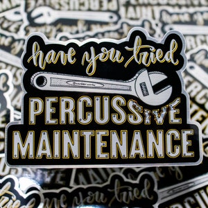 May include: A black and silver sticker with the text "have you tried PERCUSSIVE MAINTENANCE" in a stylized font. The design includes a wrench illustration and yellow accents. The sticker has a glossy finish.