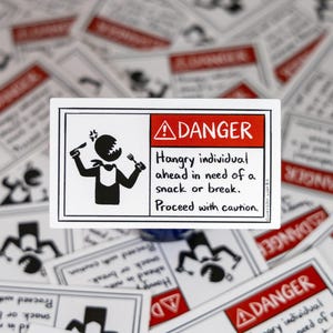 May include: A rectangular sticker with a black and white illustration of a person holding a knife and fork, with the text "DANGER Hangry individual ahead in need of a snack or break. Proceed with caution."