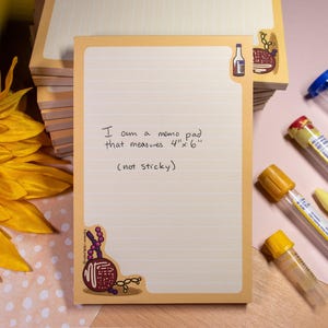 Peut inclure: Un bloc-notes avec un fond crème et une bordure orange clair. Le bloc-notes mesure 10 x 15 cm et comporte des lignes pour écrire. Le texte sur le bloc-notes indique : « I am a memo pad that measures 4" x 6" (not sticky). » Des illustrations décoratives sont dans les coins.