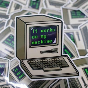 以下が含まれることがあります： 「It works on my machine」という緑色の文字が表示された画面を持つレトロなコンピューターのステッカー。ステッカーは白い縁取りとキーボードが付いています。背景には同様のステッカーが多数あります。