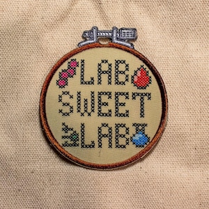 May include: A round, cross-stitched patch with the words "LAB SWEET LAB" in black thread. The patch features small embroidered images of a pink cluster, a red drop, a green flask, and a blue vial. The patch is set in a wooden embroidery hoop.