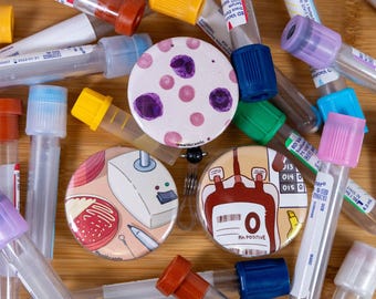 May include: An overhead shot features several medical-themed pinback buttons and test tubes. The buttons display illustrations of blood cells, blood bags, and lab equipment. The test tubes have colored caps, including red, blue, yellow, and purple.