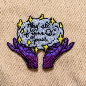 Puede incluir: Parche bordado con una nube azul claro y el texto "May all of your QC pass" en escritura negra. La nube está rodeada de estrellas amarillas y sostenida por dos manos moradas.