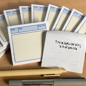 May include: A collection of square notepads with a blue border and grid pattern, labeled "Date" and "Title." A yellow pen and a gray marker are placed below the notepads. Some notepads are stacked, and one has handwritten text.