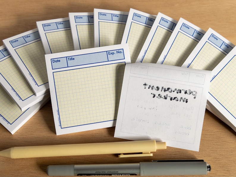May include: A collection of square notepads with a blue border and grid pattern, labeled "Date" and "Title." A yellow pen and a gray marker are placed below the notepads. Some notepads are stacked, and one has handwritten text.