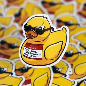 May include: A yellow rubber duck sticker wearing black sunglasses and a name tag that reads "Hello my name is OFFICIAL DEBUGGING ASSISTANT". The sticker has a white border and is surrounded by other similar stickers.