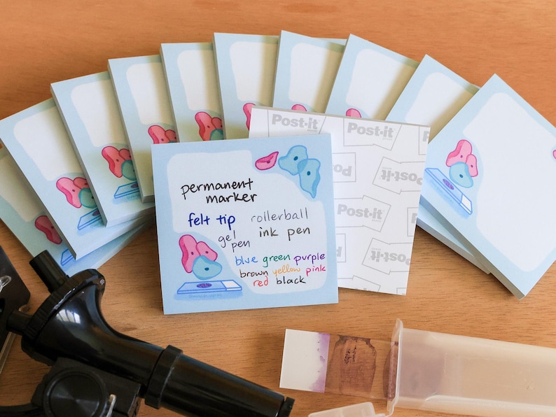 May include: A collection of light blue Post-it notes with a cartoon design, some stacked and some spread out. One note has handwritten text listing pen types and colors. A black instrument and a clear container are also visible.