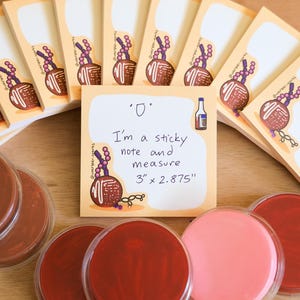 May include: A stack of peach-colored sticky notes featuring a brown candy apple illustration and the text 'I'm a sticky note and measure 3" x 2.875"'. Several round, clear containers filled with various shades of red and pink pigment are arranged below.