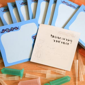 May include: A stack of light blue sticky notes with a DNA strand design border. The notes are scattered with various lab supplies, including green and orange syringes and clear pipette tips. One note has text.