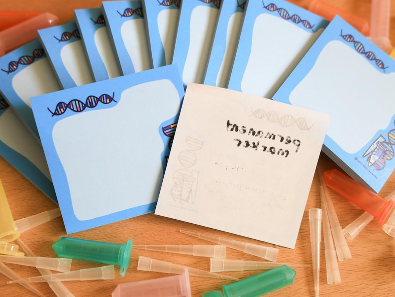 May include: A stack of light blue sticky notes with a DNA strand design border. The notes are scattered with various lab supplies, including green and orange syringes and clear pipette tips. One note has text.