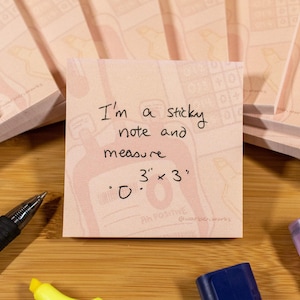 Peut inclure: Un post-it rose avec du texte manuscrit qui dit : « Je suis un post-it et mesure 7,6 cm x 7,6 cm ». La note est entourée d'autres blocs-notes et de fournitures d'écriture. L'arrière-plan est une surface en bois.