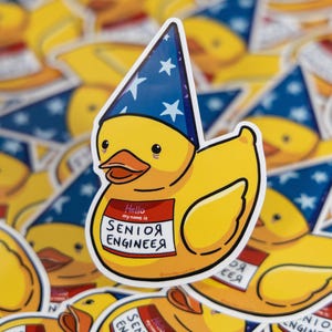 May include: A yellow duck sticker wearing a blue hat with white stars. The duck has a name tag that reads "Hello my name is SENIOR ENGINEER". The sticker is surrounded by other similar stickers.