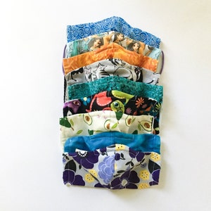 May include: A stack of 10 colorful fabric face masks with various patterns, including floral, geometric, and cartoon characters. The masks are all made of cotton and have elastic ear loops.