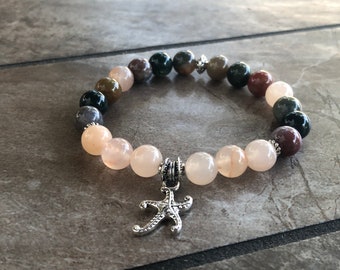 Puede incluir: Un brazalete de cuentas con una mezcla de piedras marrones, verdes y rosas. El brazalete tiene un dije de plata en forma de estrella de mar.