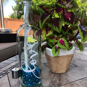 May include: A repurposed glass bottle oil lamp with the word "love" in white script. The bottle is filled with blue glass marbles and has a silver metal wick holder and a silver metal cap. A potted plant with green and burgundy leaves sits nearby.