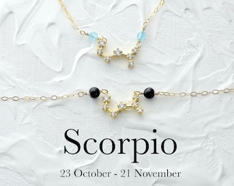 May include: A gold necklace and bracelet with a constellation design representing the zodiac sign Scorpio. The necklace has light blue beads and the bracelet has black beads. The text "Scorpio" and "23 October - 21 November" is printed below the jewelry.