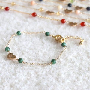 May include: A gold chain bracelet featuring small green beads and a single flat gold heart charm. The bracelet includes a lobster claw clasp and an extender chain. The design is delicate, with the metal links and beads spaced evenly throughout.