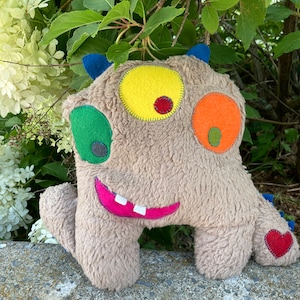 May include: A brown plush toy monster with a smiling mouth, three eyes, and a red heart on its tail. The eyes are yellow, green, and orange with black pupils.
