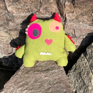 May include: A green felt monster toy with pink and orange eyes, a pink heart, and a white jagged tooth. The monster is sitting on a rock.