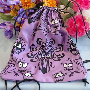 May include: A lavender drawstring backpack with a black, repeating pattern of stylized faces. The bag has black drawstrings and is set against a backdrop of colorful flowers. The design is reminiscent of a haunted house.