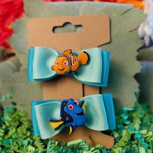 May include: Two light blue ribbon hair bows attached to a brown cardboard display card. The top bow features an orange and white clownfish character, while the bottom bow features a blue and yellow tang fish character with large eyes.