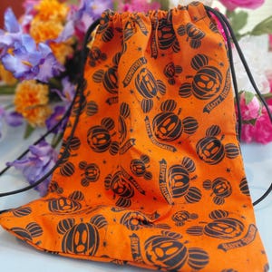 May include: Orange drawstring bag with a Halloween-themed pattern. The bag features a repeating design of black Mickey Mouse-shaped pumpkins and the text "Happy Halloween". The bag is made of fabric and has black drawstrings.