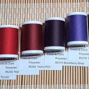 May include: A collection of spools of Coats & Clark polyester thread in various colors, including white, fuchsia, red, burgundy, purple, teal, and green. Each spool has a white base and a label with the thread's color name and number.