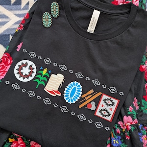 May include: A black t-shirt featuring a graphic print of a circular medallion, corn, boots, a turquoise-colored oval, tools, and a rectangular pattern, accented by two turquoise-colored oval earrings and a black floral-patterned skirt.