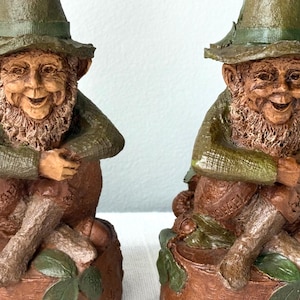 May include: Two identical gnome figurines, each wearing a green hat and green long-sleeved shirt, sitting on a brown tree stump base. The gnomes have textured brown beards, smiling faces, and crossed legs. The name Michael is inscribed on the base.