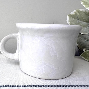 May include: A white ceramic mug with a subtle, mottled, cloud-like texture across its surface. The mug features a wide, rounded body, a thick rim, and a sturdy, curved handle on the left side. The finish has a soft, glossy sheen throughout.