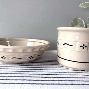 May include: A cream-colored ceramic bowl and a matching cylindrical canister, both featuring a textured basket-weave pattern around the exterior. Each piece is decorated with small, dark green floral accents and thin horizontal green lines.