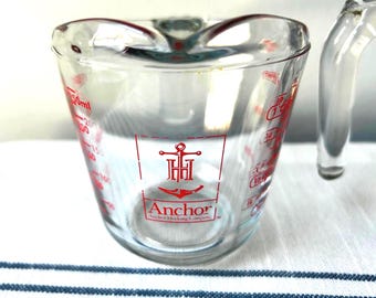 May include: A clear glass measuring cup with a red Anchor Hocking Company logo and anchor symbol printed on the front. The cup features red measurement markings in milliliters up to 250ml and imperial units including cups and fractions of a cup.