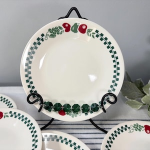 May include: A round, off-white ceramic plate featuring a decorative border of green checkered patterns, red apples, and green grapes. The plate is displayed on a black metal stand, with additional matching plates visible in the foreground.