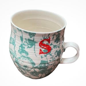 May include: White ceramic mug with a teal floral pattern and a red letter "S". The mug has a curved shape and a white handle. The interior of the mug is white.