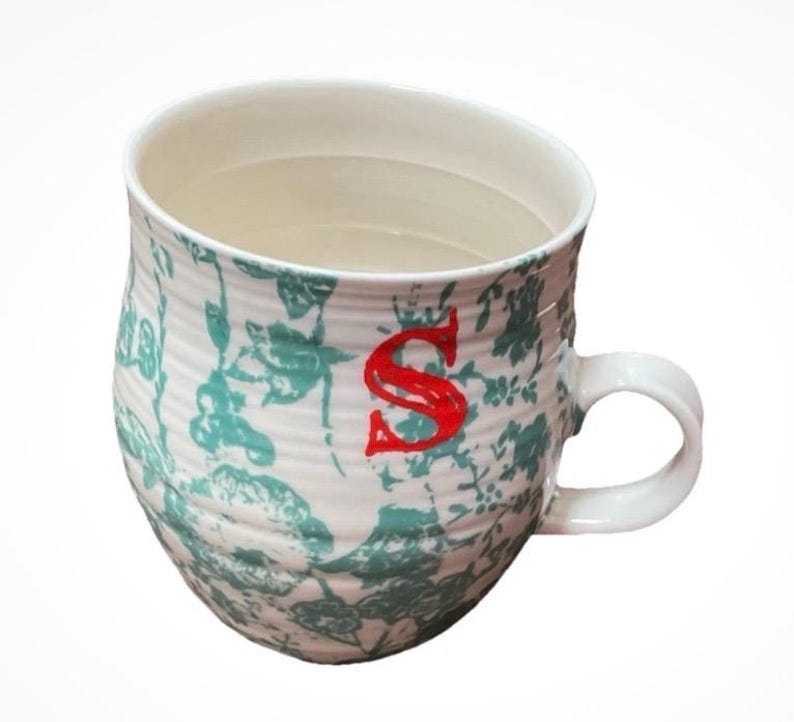 May include: White ceramic mug with a teal floral pattern and a red letter "S". The mug has a curved shape and a white handle. The interior of the mug is white.