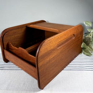 May include: A wooden bread box with a roll-top lid and a pull-out drawer. The box is made of a warm-toned wood and has a curved design. The drawer has dividers. The bread box is sitting on a striped surface.