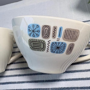 May include: Two off-white ceramic mugs featuring a repeating pattern of blue and brown geometric shapes, including starbursts, rectangles, and zig-zag lines. Each mug has a curved handle and a smooth, glossy finish on the exterior surface.